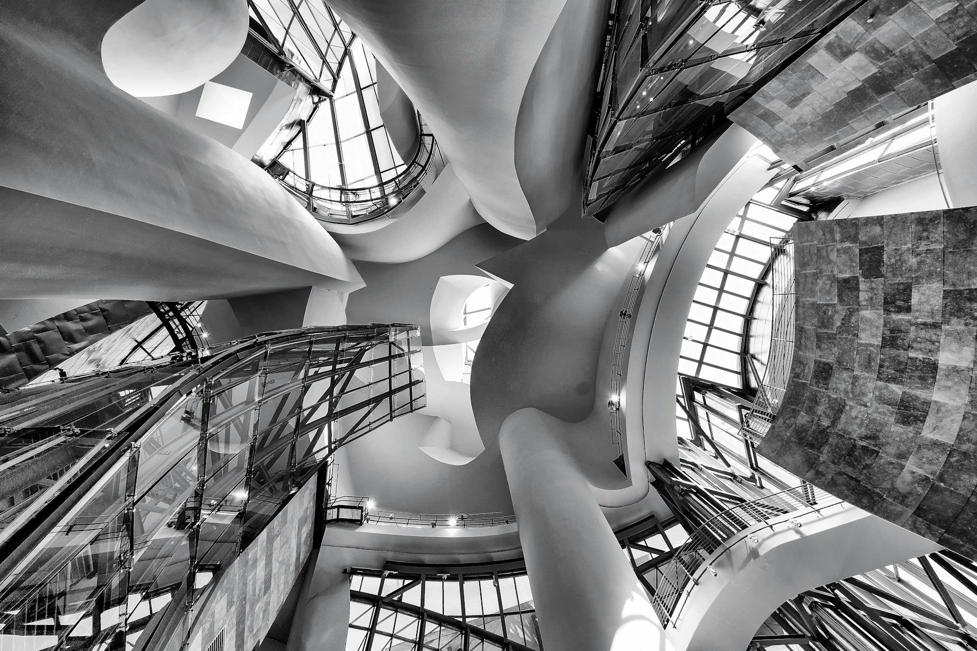 An intricate architectural interior featuring undulating curves, glass panels, and a play of light and shadows. The perspective is angled upward, capturing the dynamic and flowing design elements that create a sense of movement and modernism.