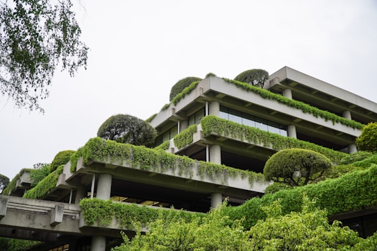 A multi-story building draped in lush green vines and shrubbery offers a harmonious blend of architecture and nature. The structure features concrete terraces and large windows, with trees and plants adorning the levels, creating a vertical garden effect.