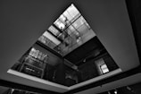A black and white architectural photograph captures the interior of a building viewed from below. The image reveals multiple floors with visible metal railings and glass panels, leading up to a skylight. The geometric composition and play of light and shadow accentuate the linear and angular elements of the architecture.