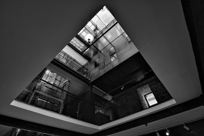 A black and white architectural photograph captures the interior of a building viewed from below. The image reveals multiple floors with visible metal railings and glass panels, leading up to a skylight. The geometric composition and play of light and shadow accentuate the linear and angular elements of the architecture.