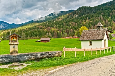 A small chapel with a wooden roof is surrounded by a lush green field and backed by dense forested mountains. A stone pillar with a cross and a sheltered niche, possibly for religious offerings, stands in the foreground. Several wooden barns or sheds are dispersed across the field, and cloudy skies loom overhead.