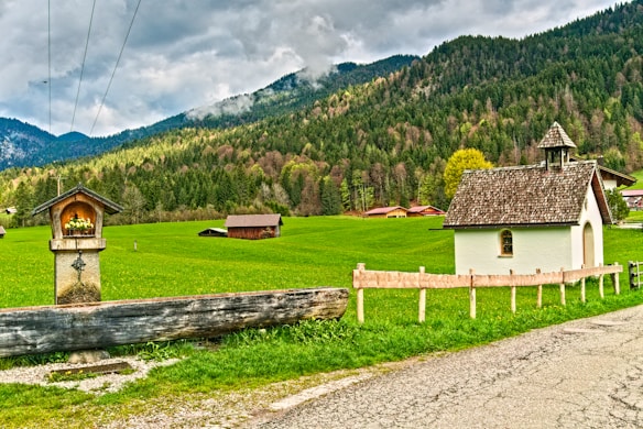 A small chapel with a wooden roof is surrounded by a lush green field and backed by dense forested mountains. A stone pillar with a cross and a sheltered niche, possibly for religious offerings, stands in the foreground. Several wooden barns or sheds are dispersed across the field, and cloudy skies loom overhead.