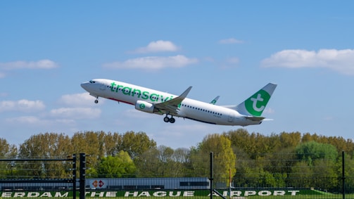 A passenger airplane is taking off from an airport, captured against a backdrop of green trees and a blue sky with a few scattered clouds. The plane has visible branding in green and white on its body.