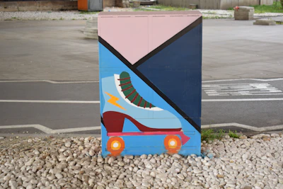 A painted utility box featuring a large graphic of a roller skate with vibrant colors. The design incorporates geometric shapes and contrasts sharply against the urban setting, with a ground surface covered in small pebbles and a paved road or walkway nearby.