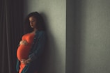 A pregnant woman stands against a patterned wall, gently cradling her belly with both hands. She wears a bright orange dress and a denim jacket. The lighting is soft and subdued, creating a calm and introspective atmosphere.