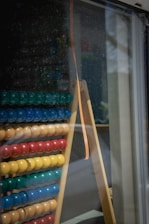 An abacus with rows of colored beads is leaning against a glass window. The beads are arranged in groups of different colors including green, blue, yellow, and red. The abacus is placed on a wooden stand with a reflective surface showing outdoor elements.