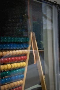 An abacus with rows of colored beads is leaning against a glass window. The beads are arranged in groups of different colors including green, blue, yellow, and red. The abacus is placed on a wooden stand with a reflective surface showing outdoor elements.