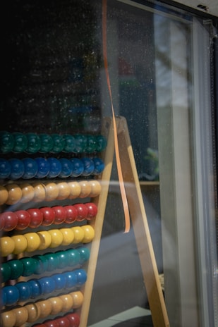 An abacus with rows of colored beads is leaning against a glass window. The beads are arranged in groups of different colors including green, blue, yellow, and red. The abacus is placed on a wooden stand with a reflective surface showing outdoor elements.
