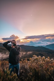 A person is taking photographs with a camera in a mountainous landscape during sunset or sunrise. The sky is filled with scattered clouds and a gradient of warm colors, with vibrant foliage in the foreground.