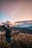 A person is taking photographs with a camera in a mountainous landscape during sunset or sunrise. The sky is filled with scattered clouds and a gradient of warm colors, with vibrant foliage in the foreground.