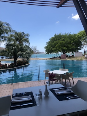 A picturesque outdoor dining setting by a pool, with palm trees and lush greenery in the background. The table is elegantly set with white napkins, cutlery, and a single flower in a vase. The sky is clear and blue, reflecting tranquility.