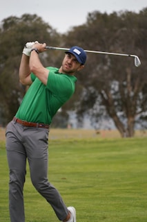 A man in a green shirt and gray pants is playing golf on a grassy course. He is completing a golf swing with his eyes focused on the ball. He is wearing a blue cap and white shoes, and there are trees in the background.