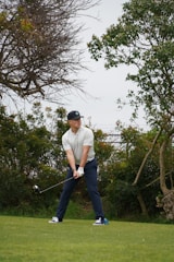 A person is standing on a grassy field, holding a golf club in a ready-to-swing position. The individual is wearing a cap, a light-colored shirt, dark pants, and sports shoes. Surrounding the player are lush green bushes and trees, indicating a natural setting possibly on a golf course.