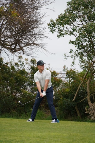 A person is standing on a grassy field, holding a golf club in a ready-to-swing position. The individual is wearing a cap, a light-colored shirt, dark pants, and sports shoes. Surrounding the player are lush green bushes and trees, indicating a natural setting possibly on a golf course.