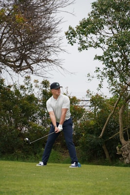 A person is standing on a grassy field, holding a golf club in a ready-to-swing position. The individual is wearing a cap, a light-colored shirt, dark pants, and sports shoes. Surrounding the player are lush green bushes and trees, indicating a natural setting possibly on a golf course.