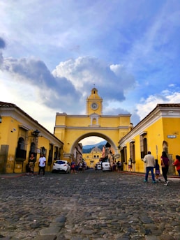 A cobblestone street leads to a yellow arch with a clock, flanked by vibrant colonial buildings against a backdrop of a blue sky with scattered clouds. People are walking and cars are parked along the road, conveying a bustling yet historical atmosphere.