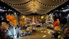 A lavishly decorated event space featuring elegant drapery and numerous floral arrangements. Warm yellow and orange tones dominate the decor, with large, illuminated flowers and delicate, hanging lights creating a whimsical ambiance. The ceiling is adorned with chandeliers and strings of lights, while the path is lined with intricate floral designs.