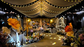 A lavishly decorated event space featuring elegant drapery and numerous floral arrangements. Warm yellow and orange tones dominate the decor, with large, illuminated flowers and delicate, hanging lights creating a whimsical ambiance. The ceiling is adorned with chandeliers and strings of lights, while the path is lined with intricate floral designs.