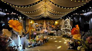 A lavishly decorated event space featuring elegant drapery and numerous floral arrangements. Warm yellow and orange tones dominate the decor, with large, illuminated flowers and delicate, hanging lights creating a whimsical ambiance. The ceiling is adorned with chandeliers and strings of lights, while the path is lined with intricate floral designs.