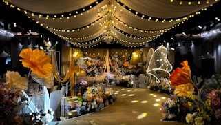 A lavishly decorated event space featuring elegant drapery and numerous floral arrangements. Warm yellow and orange tones dominate the decor, with large, illuminated flowers and delicate, hanging lights creating a whimsical ambiance. The ceiling is adorned with chandeliers and strings of lights, while the path is lined with intricate floral designs.