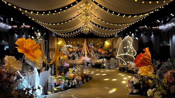 A lavishly decorated event space featuring elegant drapery and numerous floral arrangements. Warm yellow and orange tones dominate the decor, with large, illuminated flowers and delicate, hanging lights creating a whimsical ambiance. The ceiling is adorned with chandeliers and strings of lights, while the path is lined with intricate floral designs.