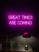 A neon sign with the words 'GREAT TIMES ARE COMING' in bright pink hangs on a dark wall. Below it, a sleek black desk lamp sits on a reflective surface, alongside a small green plant and a geometric glass ornament.