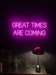 A neon sign with the words 'GREAT TIMES ARE COMING' in bright pink hangs on a dark wall. Below it, a sleek black desk lamp sits on a reflective surface, alongside a small green plant and a geometric glass ornament.