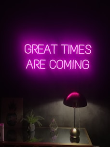 A neon sign with the words 'GREAT TIMES ARE COMING' in bright pink hangs on a dark wall. Below it, a sleek black desk lamp sits on a reflective surface, alongside a small green plant and a geometric glass ornament.