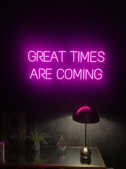 A neon sign with the words 'GREAT TIMES ARE COMING' in bright pink hangs on a dark wall. Below it, a sleek black desk lamp sits on a reflective surface, alongside a small green plant and a geometric glass ornament.