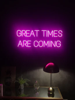 A neon sign with the words 'GREAT TIMES ARE COMING' in bright pink hangs on a dark wall. Below it, a sleek black desk lamp sits on a reflective surface, alongside a small green plant and a geometric glass ornament.