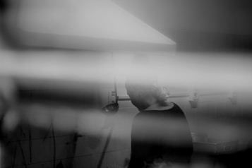 A black and white image of a person wearing a headscarf appears to be working in a kitchen environment. The figure is slightly blurred and obscured by horizontal lines, possibly from an out-of-focus foreground object or reflections. A kitchen vent hood is visible above the person.