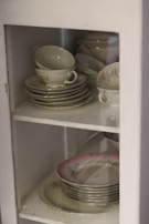A collection of faded floral china cups arranged on a lace-covered shelf.