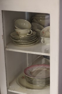 A collection of faded floral china cups arranged on a lace-covered shelf.