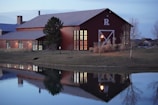 A red barn-like building with large windows is reflected in a tranquil pond surrounded by grass and sparse trees. The building has a large 'R' on its facade and a chimney on one side. Soft exterior lights illuminate the structure, suggesting early evening or dusk.