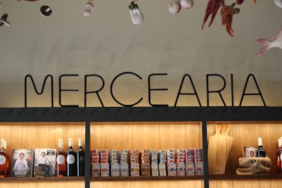 Interior display of a grocery shop featuring a prominent 'MERCEARIA' sign above wooden shelves. The shelves are stocked with bottles of wine, packaged food items like canned goods or boxes, and cutting boards. The ambiance is warm with illuminated shelving highlighting the products. Decorative items hang from the ceiling, including peppers and other hanging produce.