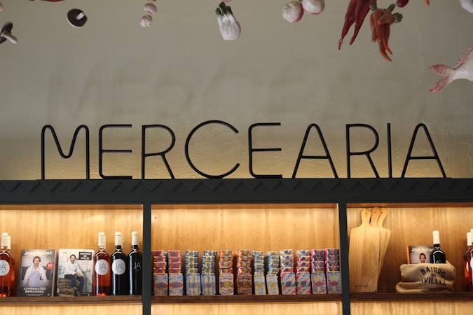 Interior display of a grocery shop featuring a prominent 'MERCEARIA' sign above wooden shelves. The shelves are stocked with bottles of wine, packaged food items like canned goods or boxes, and cutting boards. The ambiance is warm with illuminated shelving highlighting the products. Decorative items hang from the ceiling, including peppers and other hanging produce.