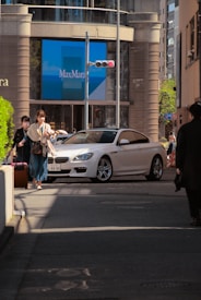 A city street scene features a white luxury car turning at an intersection. Two pedestrians are walking on the sidewalk, carrying bags. Behind them, a large storefront displays the name 'MaxMara' on a blue backdrop. Sunlight casts shadows on the street, and tall buildings surround the area.