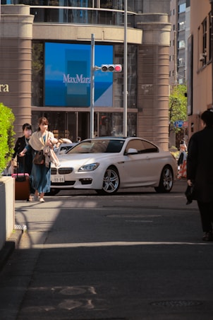 A city street scene features a white luxury car turning at an intersection. Two pedestrians are walking on the sidewalk, carrying bags. Behind them, a large storefront displays the name 'MaxMara' on a blue backdrop. Sunlight casts shadows on the street, and tall buildings surround the area.