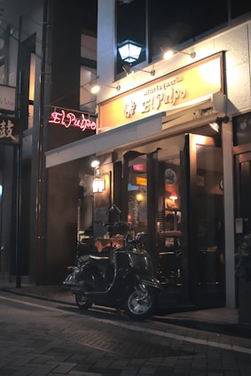 A dimly lit street scene at night featuring a small restaurant or cafe with an illuminated sign that reads 'El Pulpo'. A vintage scooter is parked on the brick-paved sidewalk in front of the entrance. Warm lights inside the establishment create a cozy and inviting atmosphere.
