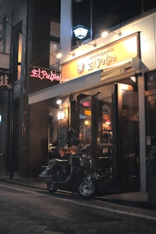 A dimly lit street scene at night featuring a small restaurant or cafe with an illuminated sign that reads 'El Pulpo'. A vintage scooter is parked on the brick-paved sidewalk in front of the entrance. Warm lights inside the establishment create a cozy and inviting atmosphere.