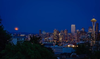 A nighttime skyline view of a city with illuminated skyscrapers and the iconic Space Needle in the foreground. A full moon rises in the background, casting a soft glow over the scene. Lush greenery is partially visible at the bottom of the image, adding contrast to the urban landscape.