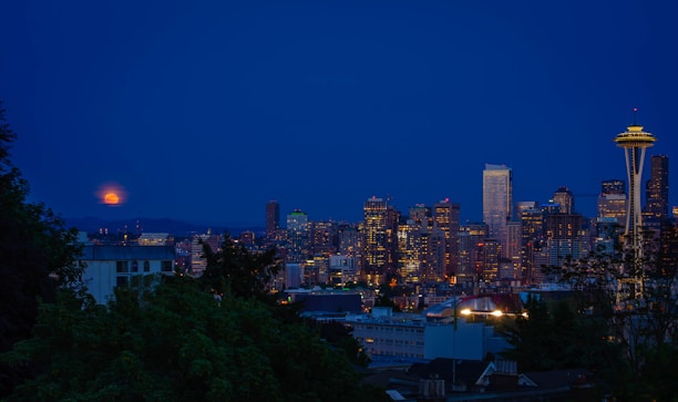 A nighttime skyline view of a city with illuminated skyscrapers and the iconic Space Needle in the foreground. A full moon rises in the background, casting a soft glow over the scene. Lush greenery is partially visible at the bottom of the image, adding contrast to the urban landscape.