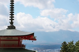 A traditional Japanese pagoda stands prominently in the foreground, with its tiered roof and architectural details visible. In the background, the snow-capped peak of Mount Fuji emerges through scattered clouds. The sky is a soft blue with patches of white clouds, and the surrounding landscape appears lush with greenery.