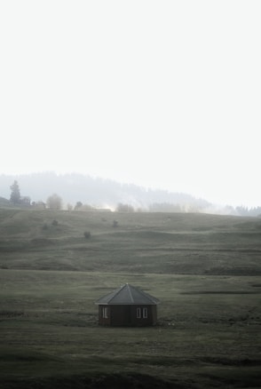 A solitary wooden hut with a green roof sits in the middle of expansive, gentle rolling hills covered in grass. The background features a hazy and soft-lit tree line with mist adding an ethereal quality, suggesting a serene and isolated countryside setting.