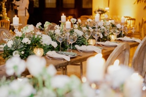 An elegantly set table with white floral arrangements and greenery cascading down the center. The table is adorned with candles and sparkling glassware, creating a warm and inviting atmosphere. The soft lighting enhances the overall sophisticated ambience.