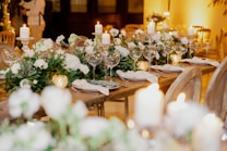 An elegantly set table with white floral arrangements and greenery cascading down the center. The table is adorned with candles and sparkling glassware, creating a warm and inviting atmosphere. The soft lighting enhances the overall sophisticated ambience.