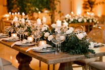 A beautifully set dining table with elegant glassware, white plates, and neatly folded napkins. The table is adorned with white roses and lush greenery, accompanied by white candles in ornate candle holders. The warm ambient lighting creates a cozy and inviting atmosphere, with blurred floral arrangements visible in the background.