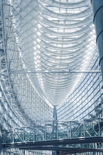 A modern architectural interior featuring a large, curved glass and steel ceiling. The design includes a series of arched structures and a complex network of metal beams. Sunlight filters through the glass, creating patterns of light and shadow on the surfaces. A suspended walkway is visible below, emphasizing the spaciousness and complexity of the structure.