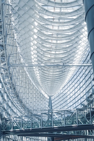 A modern architectural interior featuring a large, curved glass and steel ceiling. The design includes a series of arched structures and a complex network of metal beams. Sunlight filters through the glass, creating patterns of light and shadow on the surfaces. A suspended walkway is visible below, emphasizing the spaciousness and complexity of the structure.