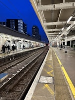 A train platform with tracks running parallel, bordered by a covered station with white beams. Several people are walking along the platform, and tall buildings can be seen in the background against a dark blue evening sky. The platform is illuminated by overhead lights, creating a contrast with the dark surroundings.